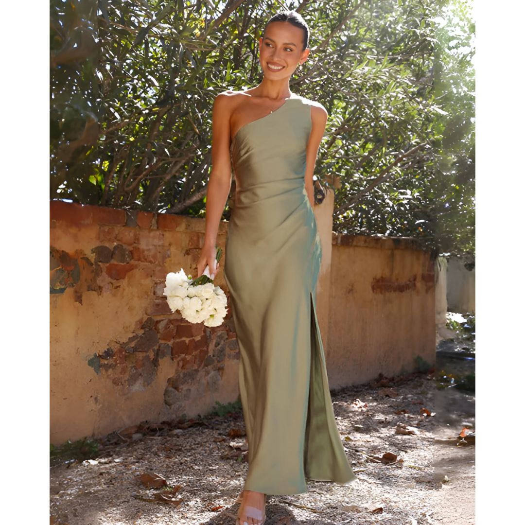 Woman in a long green dress holding flowers against a rustic wall and tree background