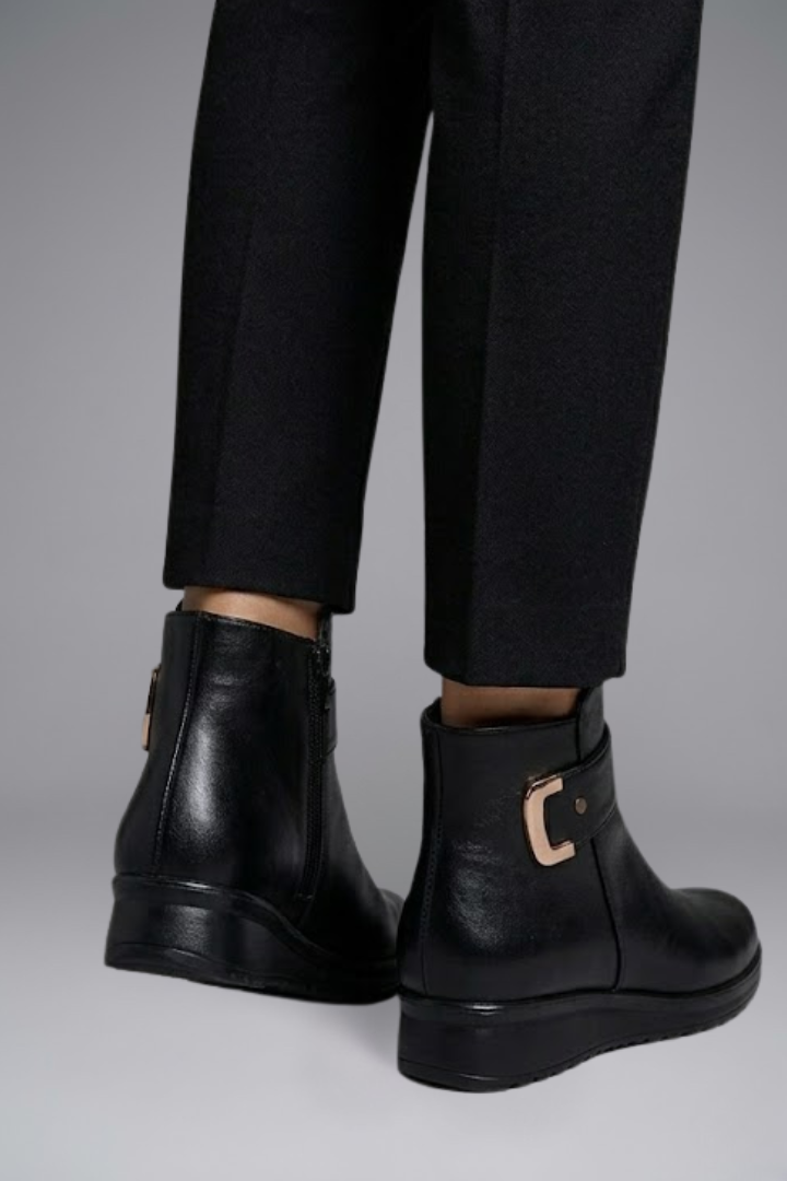 Black leather boots with a buckle detail worn with black pants on a gray background
