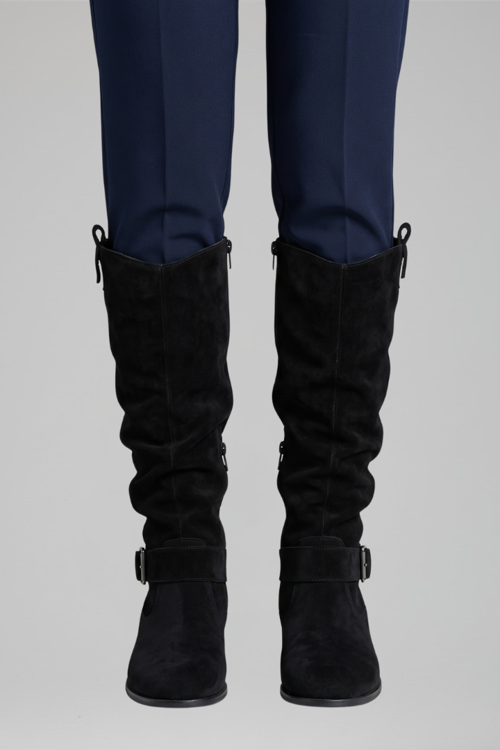 Black knee-high boots with buckles worn with navy pants on a light gray background