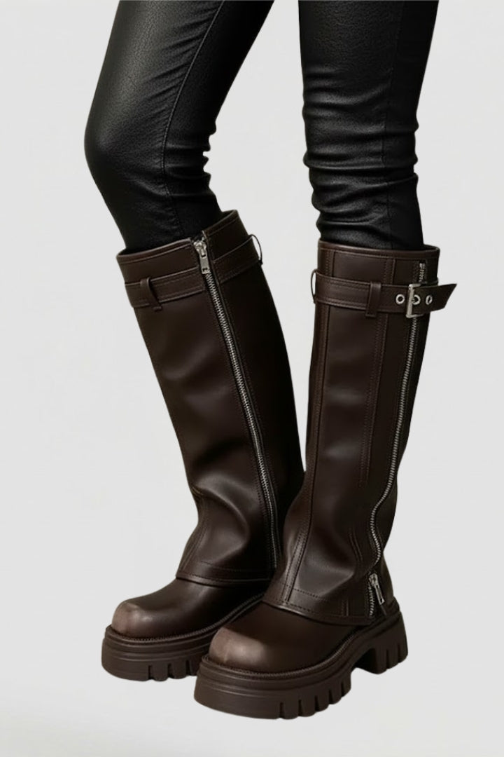 Brown knee-high boots with zippers and buckles worn with black pants on a light gray background
