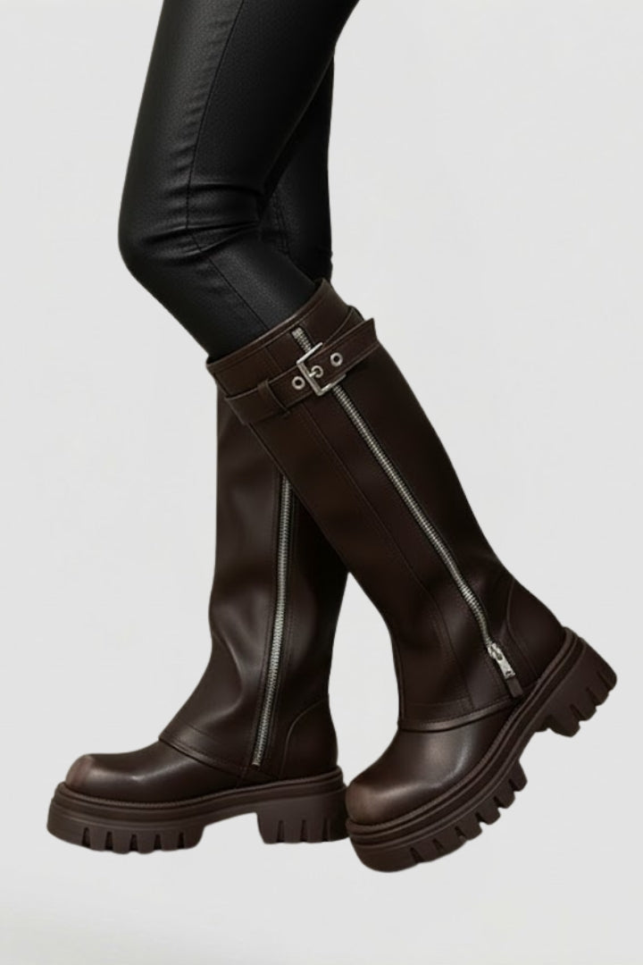 Brown knee-high boots with silver buckles worn by a person on a light gray background
