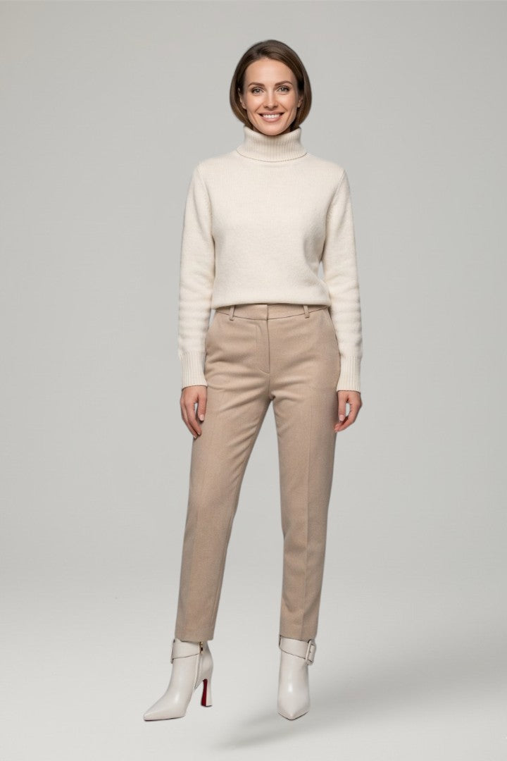 Woman wearing a cream turtleneck sweater and beige pants on a gray background