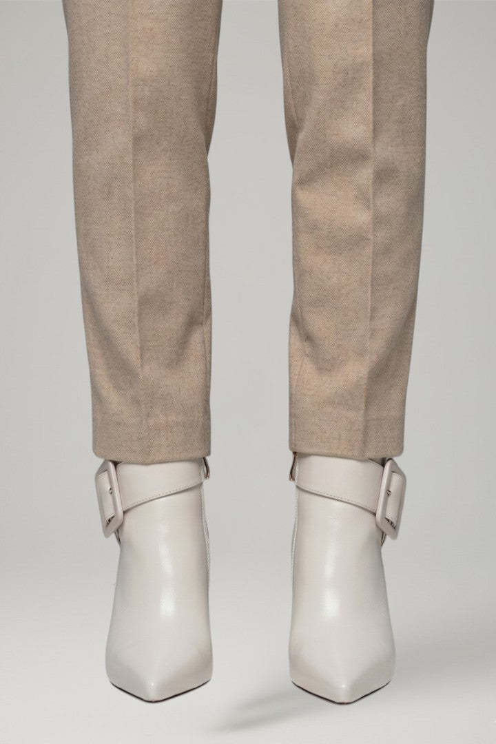 Close-up of a person's legs wearing beige trousers and off-white pointed-toe ankle boots with wraparound straps and square buckle details, set against a neutral light gray studio background.