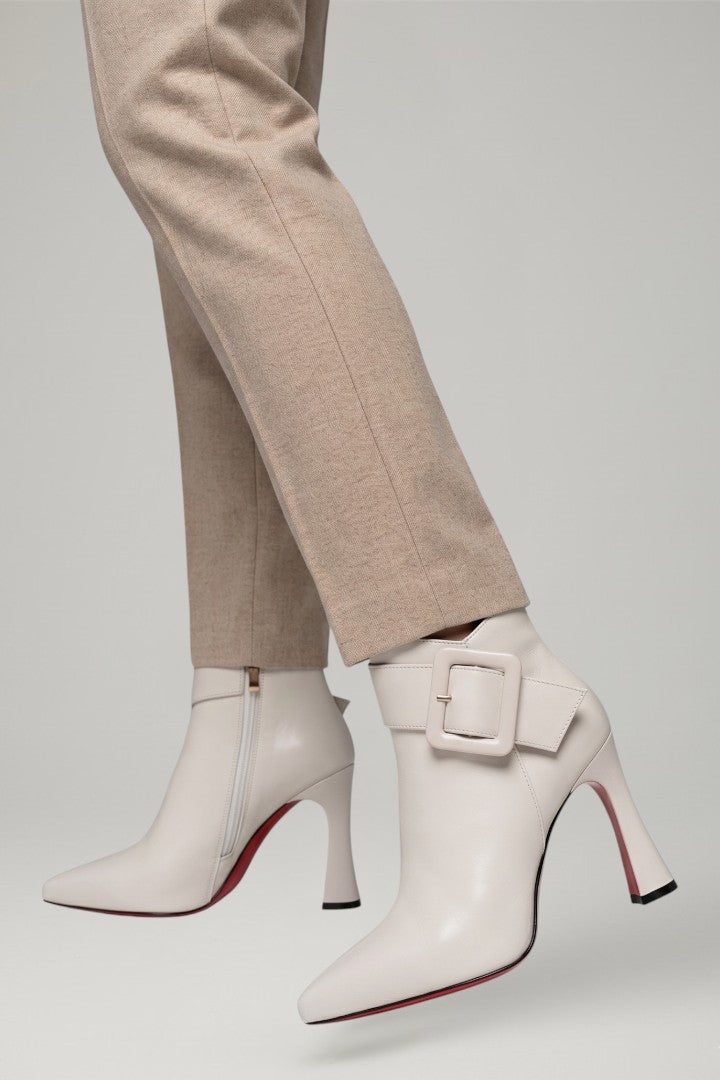 White ankle boots with a high heel and red sole, worn with beige pants on a light gray background.