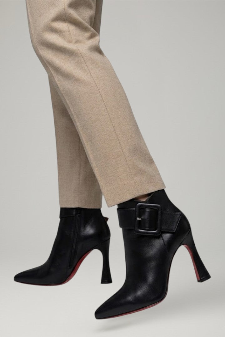 Black high-heeled boots with a buckle detail worn with beige pants on a light gray background