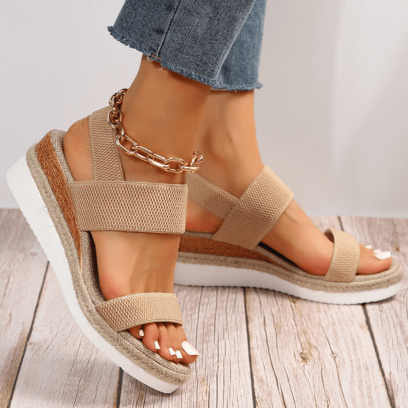 Beige wedge sandals with chain detail worn on a wooden floor.