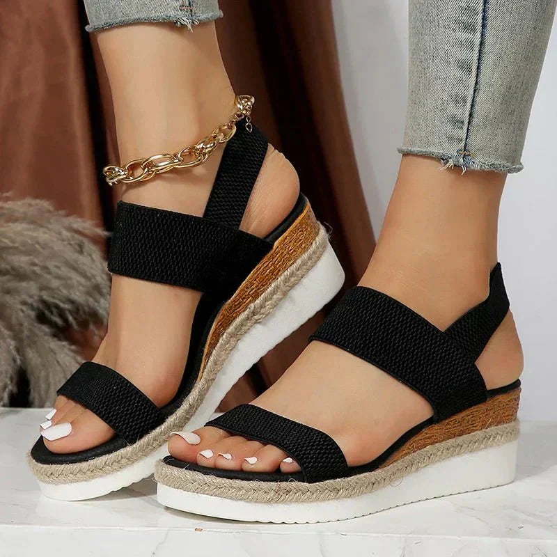 Black wedge sandals with a wooden sole worn by a person, against a neutral background.