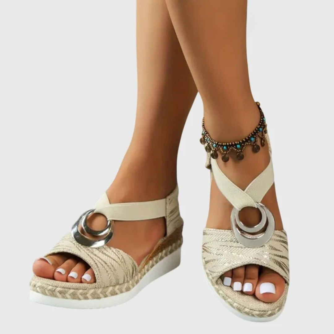 Beige sandals with metallic ring design worn by a person on a light gray background