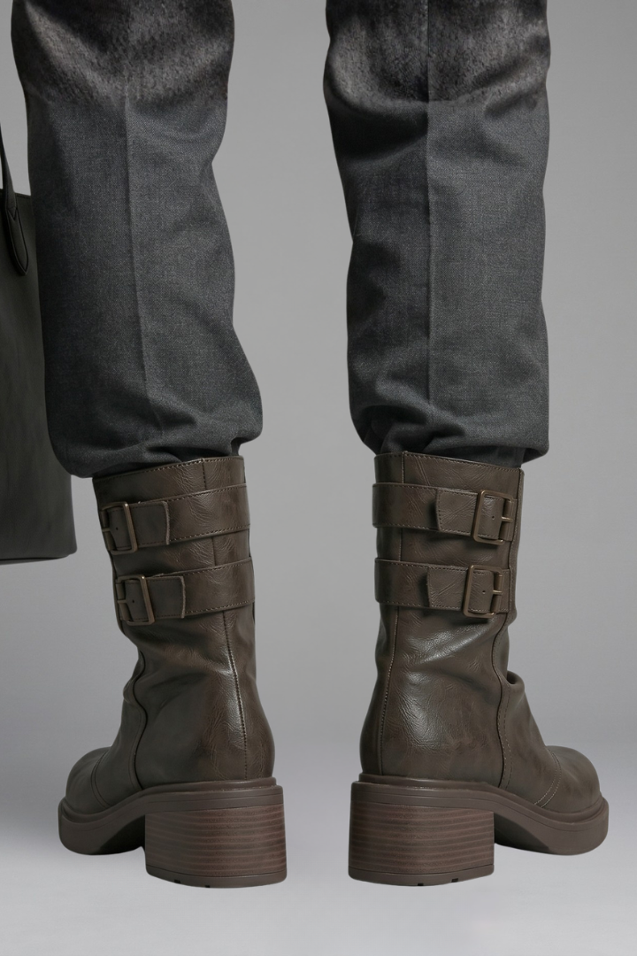 Brown leather boots with buckles worn with gray pants on a neutral background