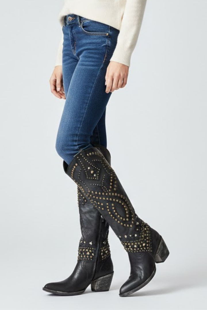 Person wearing blue jeans and black knee-high boots with gold studs on a light gray background