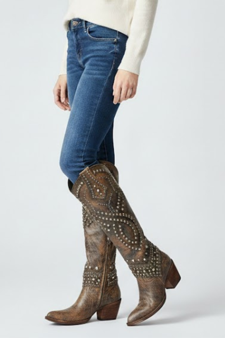 Person wearing blue jeans and brown studded knee-high boots on a light gray background