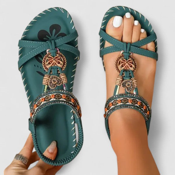 Teal sandals with decorative straps and charms held by a person on a light gray background