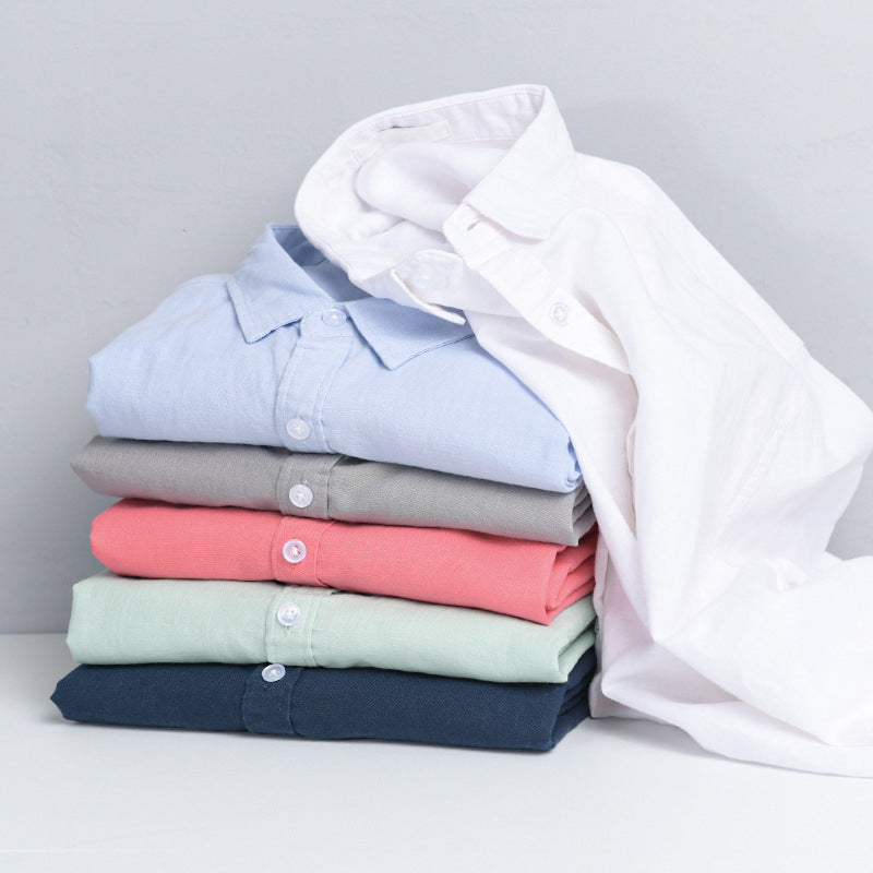 Stack of folded shirts in various colors with a white shirt leaning against them on a light gray background.