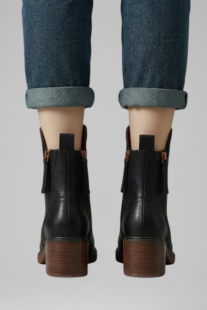 Black leather ankle boots with brown soles worn with rolled-up jeans on a light gray background