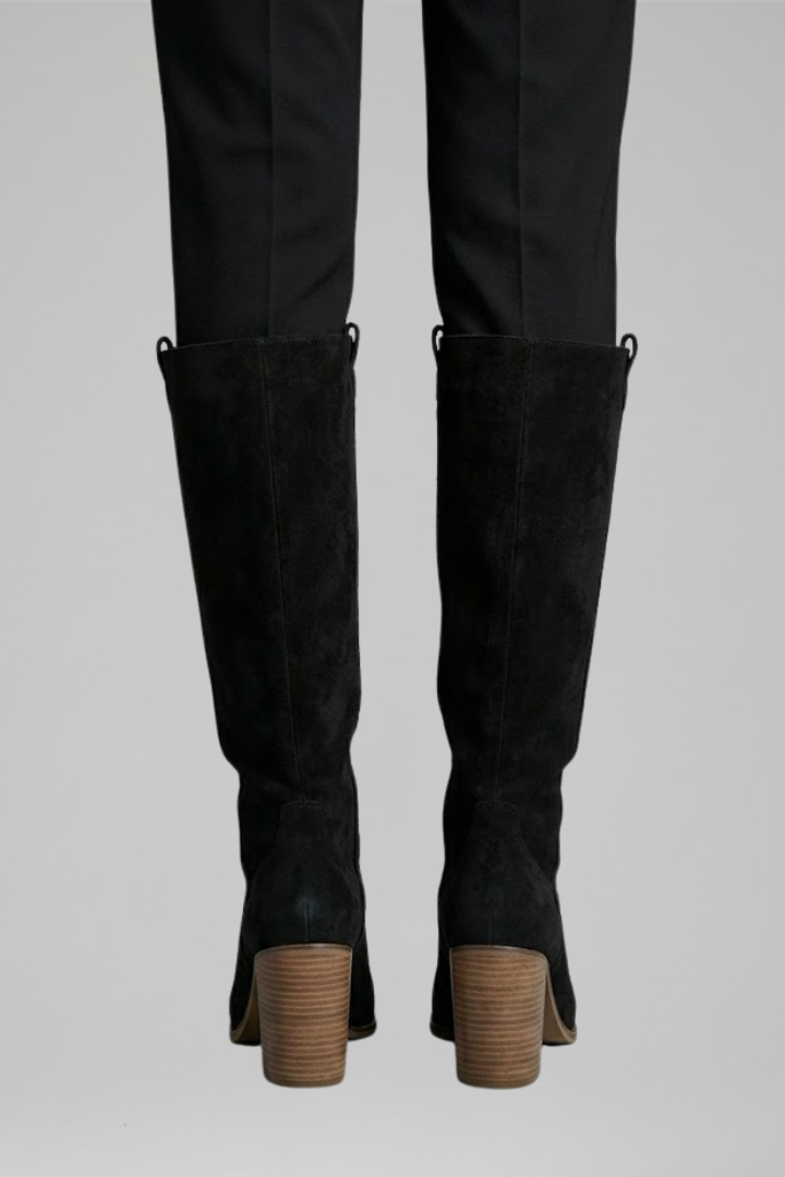 Black knee-high boots with brown soles worn with black pants on a light gray background